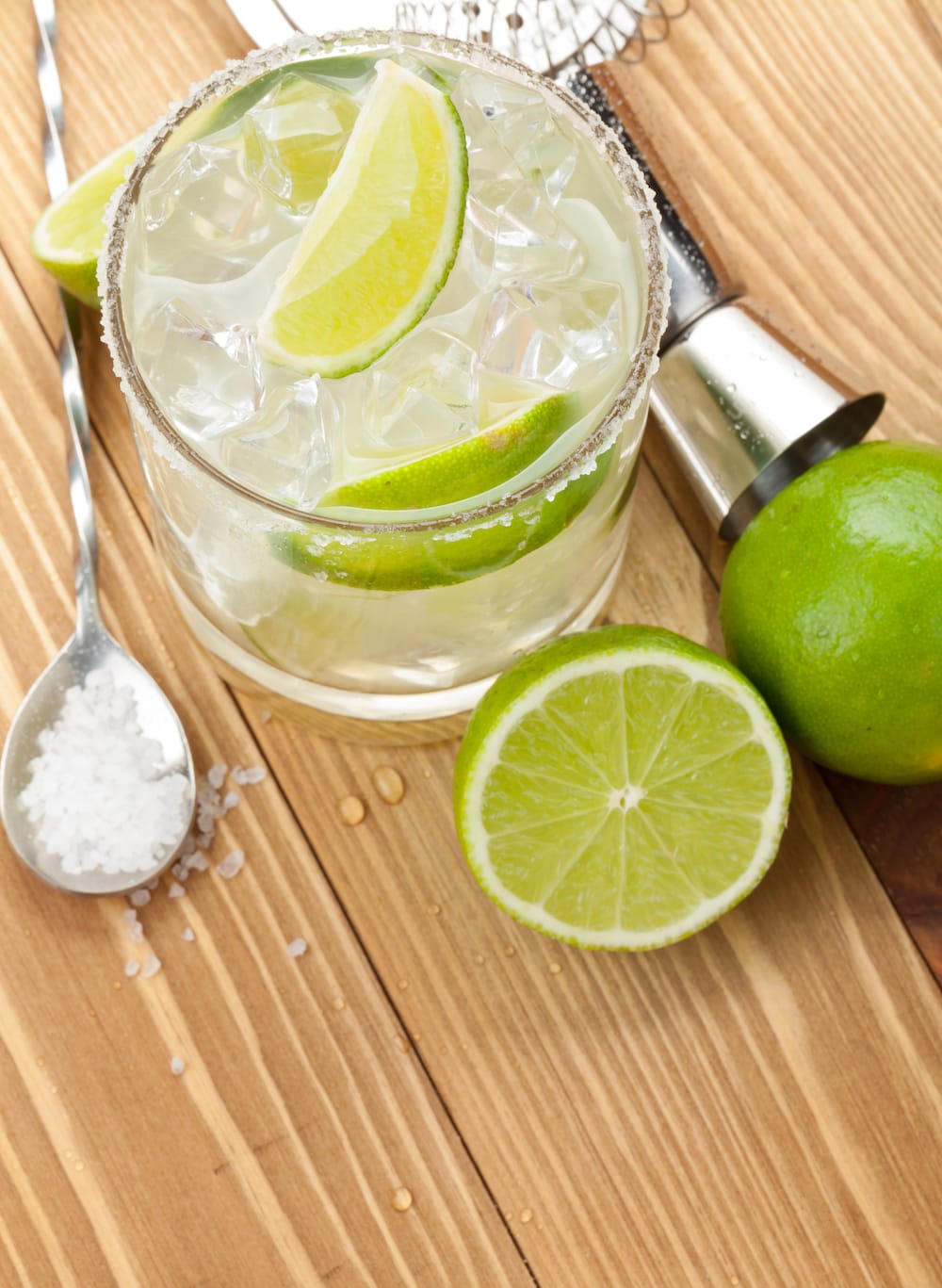 A classic margarita cocktail with drink utensils and limes on wooden table. 