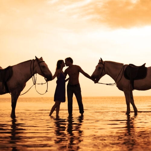 Couple horseback riding on the beach at sunset.