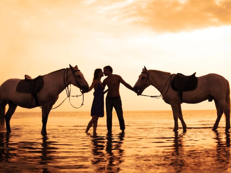 Couple horseback riding on the beach at sunset.