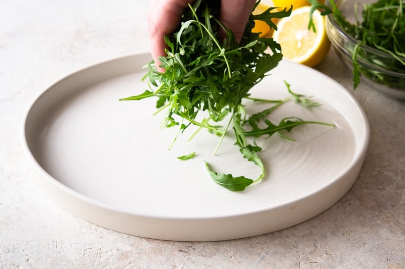 A person scattering arugula on a white platter. 