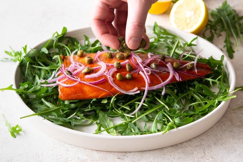 A person's hand garnishing trout with capers and slivered red onion. 