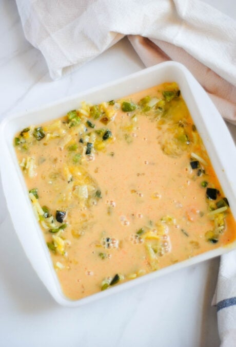 Whisked eggs being poured over a vegetarian casserole in a white dish.