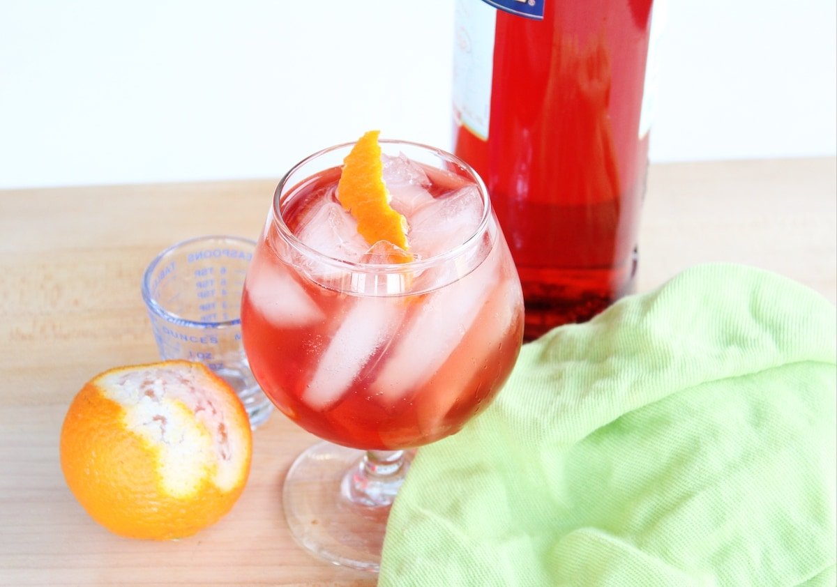 Orange, a bottle of Campari and a cocktail with a green napkin on a countertop.