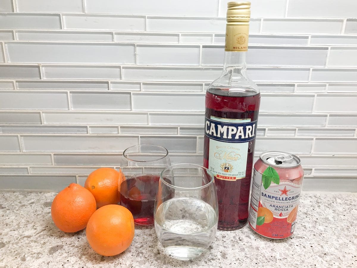 Campari, pomegranate juice, vodka, oranges and San Pellegrino sparkling drink on a countertop.