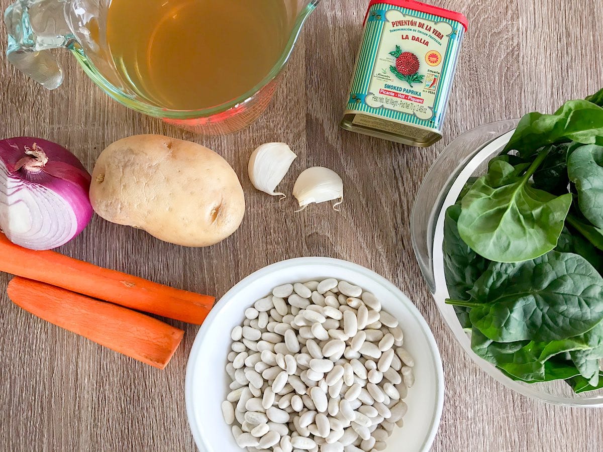 Ingredients for White Bean Soup including carrot, potato, red onion, garlic, vegetable broth, smoked paprika, baby spinach and white kidney beans on a wooden table.