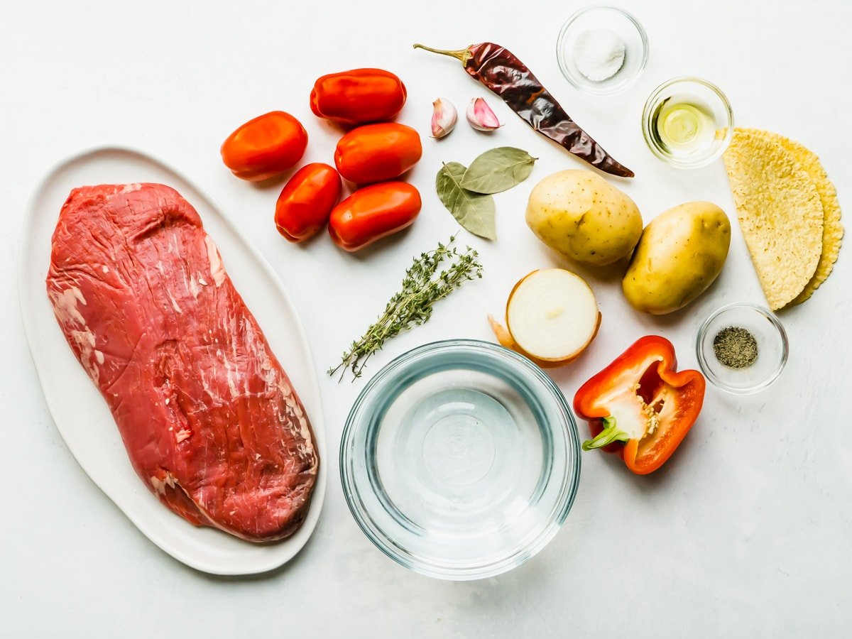 Overhead shot of ingredients for hilachas including flank steak, garlic, onion, tomatoes, red pepper, potato, chile, salt, pepper, thyme and bay leaf.