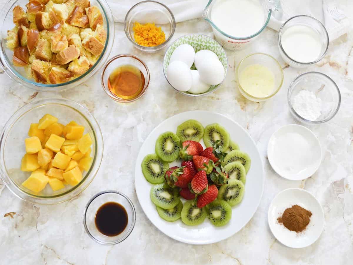 Ingredients for Baked French Toast including bread, orange zest, milk, cream, mango chunks, syrup, eggs, butter, powdered sugar, vanilla, kiwi and strawberries, granulated sugar, cinnamon.