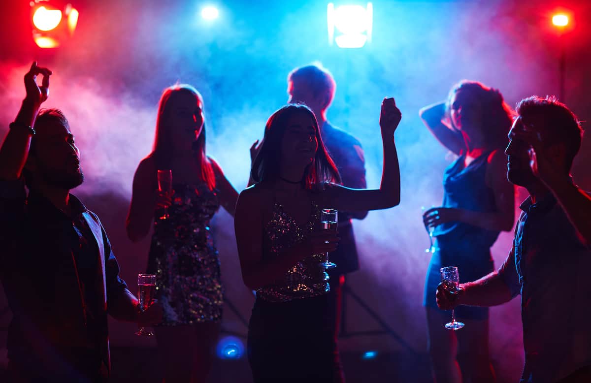 Silhouettes of young dressed-up people dancing in club smoke with glasses of champagne.