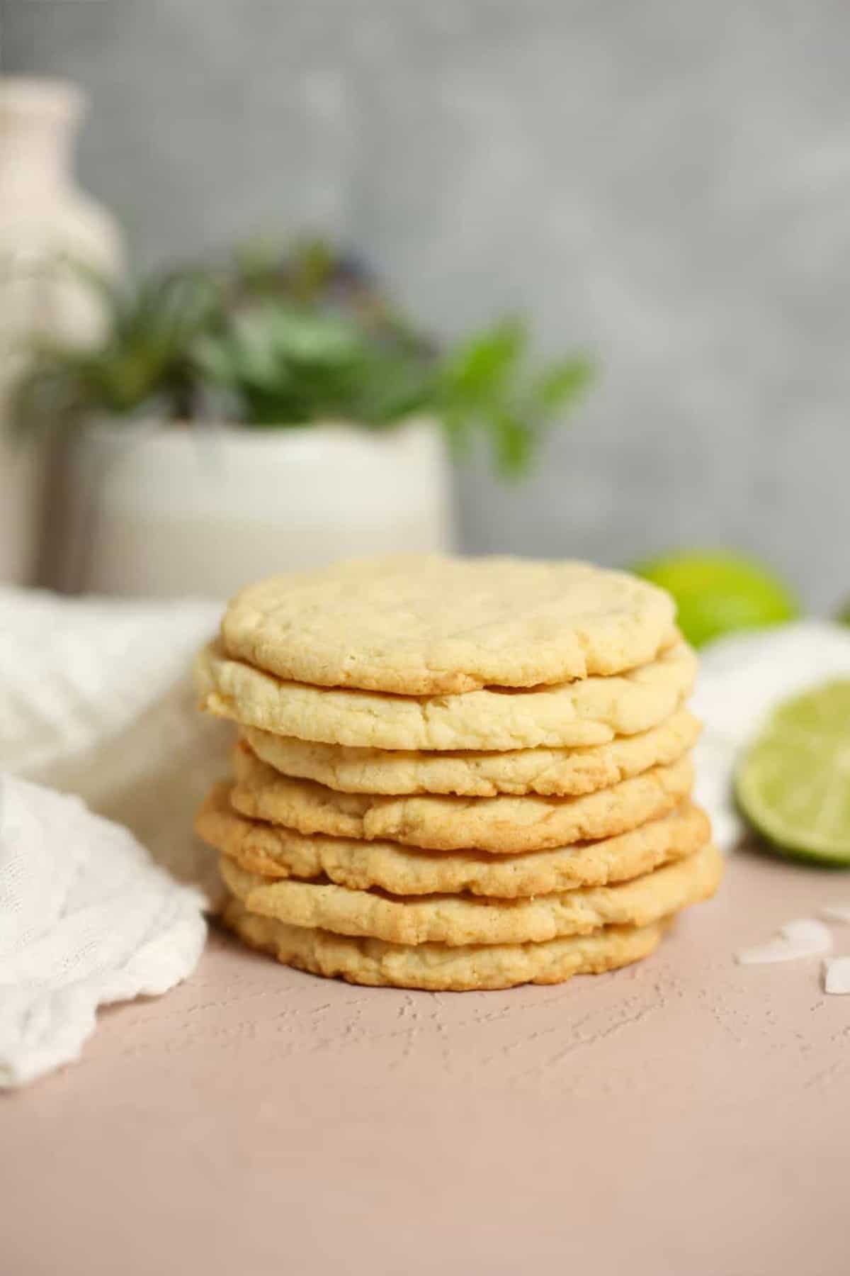 A short stack of coconut sugar cookies on a table with two flat cookies in front and limes in back.