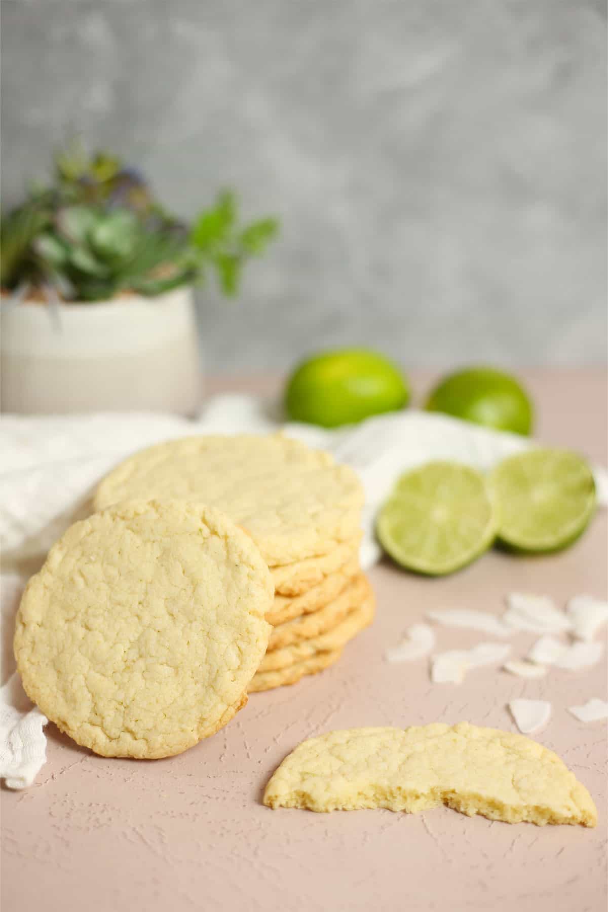 A short stack of coconut lime cookies on a table with two flat cookies in front and limes in back.