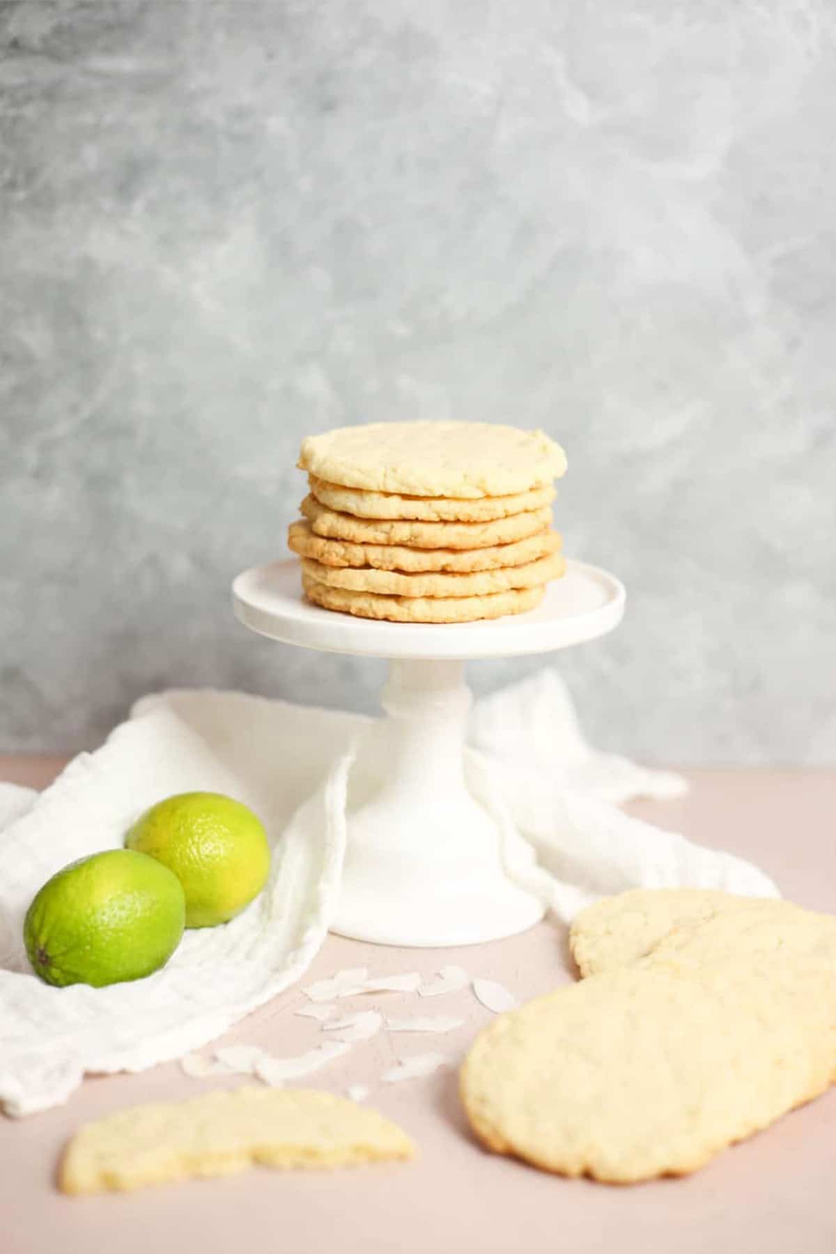 A short stack of Coconut Lime Cookies on a pedestal with two flat cookies in front and limes in back.