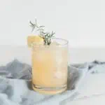An old-fashioned glass filled with ice and a chamomile cocktail garnished with rosemary and lemon.. d ice and