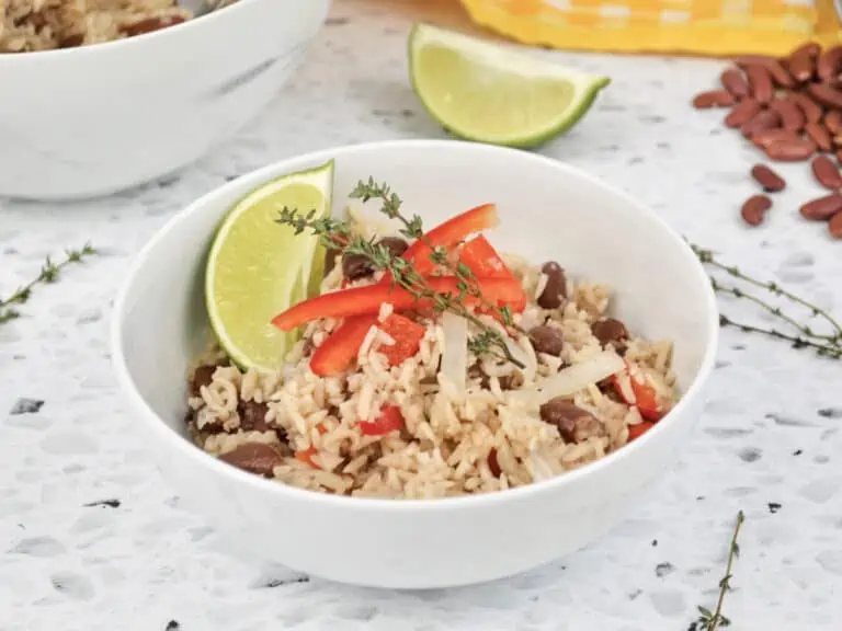 Caribbean rice and red beans in a white bowl garnished with thyme, red pepper and lime wedge.