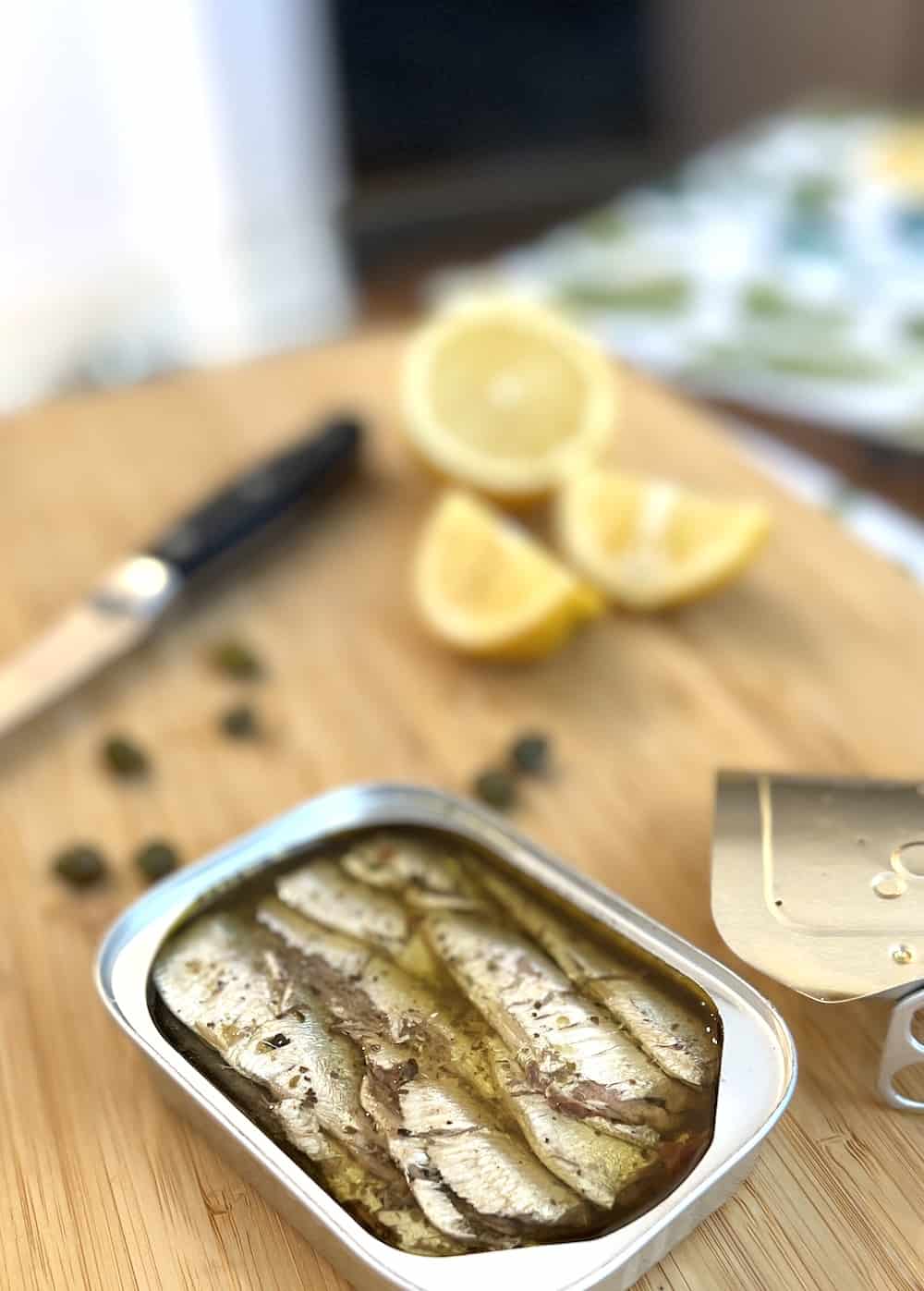 Close-up of canned sardines with a knife, lemon and capers on a board. 