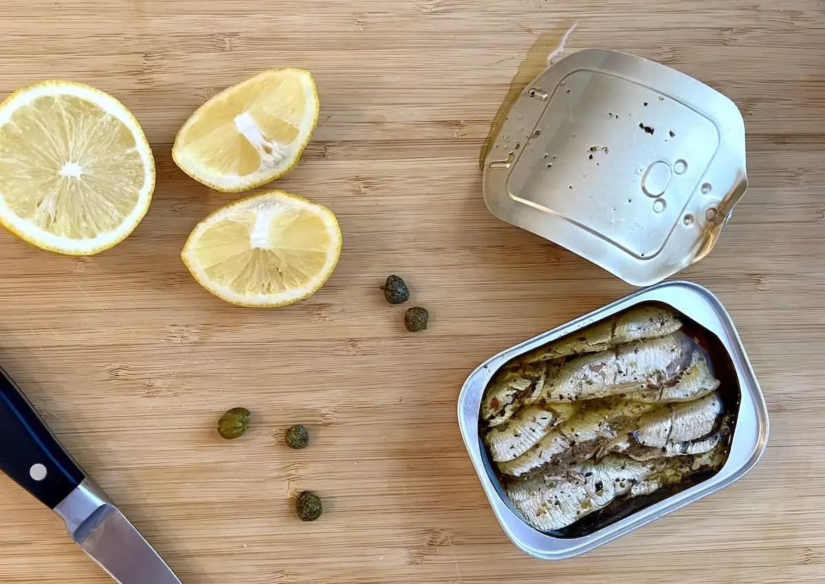 A tin of wild brisling sardines on a board with a knife, a sliced lemon and capers.