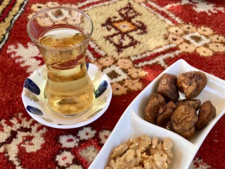 A glass of Turkish tea with nuts on a white plate on a red rug.