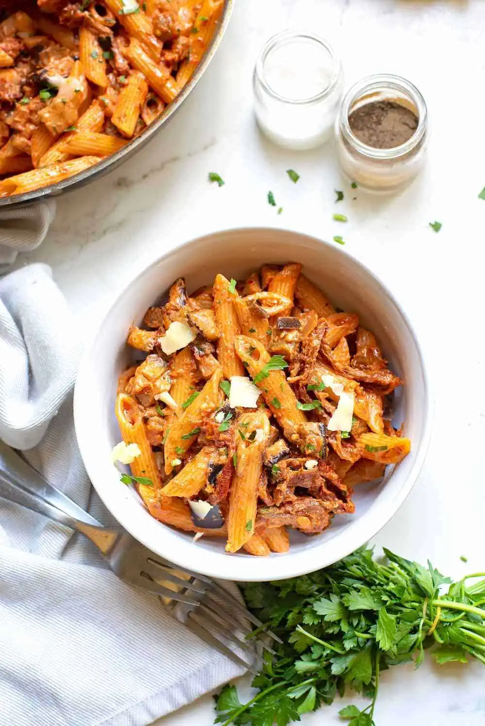 A bowl of sundried tomato pasta with eggplant on the table with parsley, salt, and pepper.