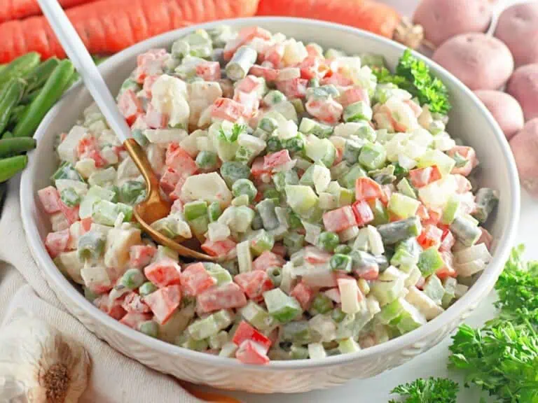 A white bowl of Ensalada Rusa (Russian Salad) surrounded by vegetables.