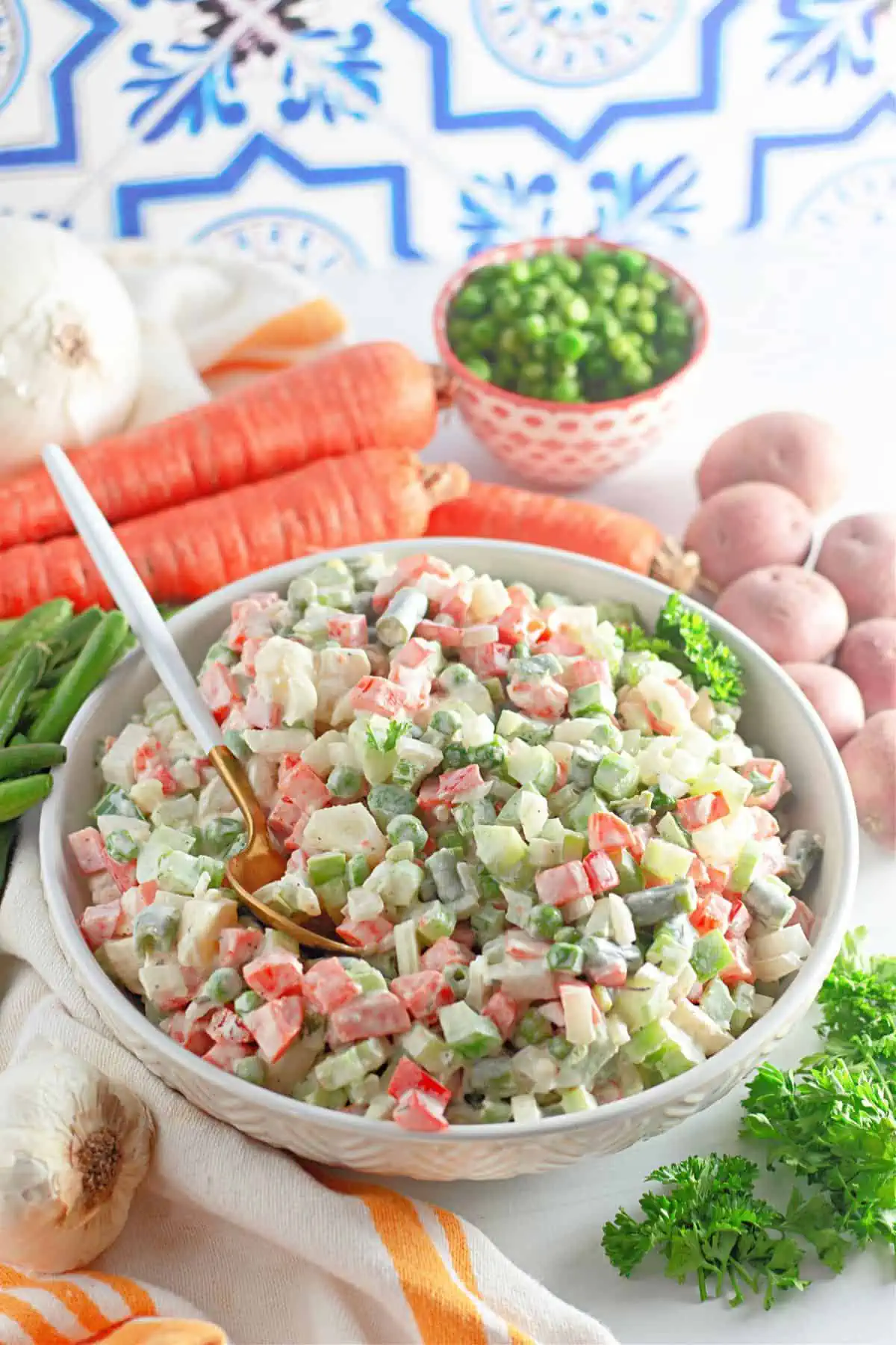 A bowl of Guatemalan Russian Salad on the table with a spoon in the bowl and vegetables in the background.