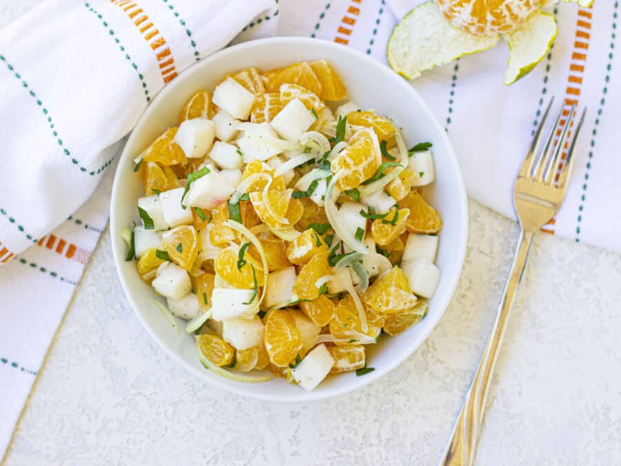 A bowl of jicama orange salad with a white tea towel in the back.