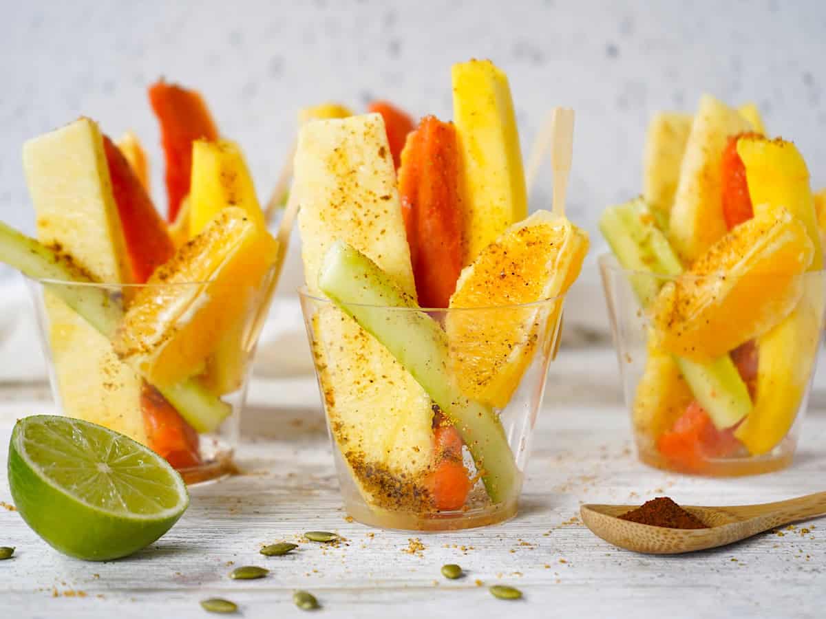 Three fruit cups sprinkled with pepitoria on a counter with a lime half to the side. 